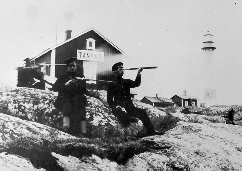 Vanha mustavalkokuva Tankarin majakkasaaresta. Kaksi miestä tarkkailee kivääreiden kanssa merelle ja yksi mies tarkkailee kaukoputkella. Kuva: K.H. Renlundin museo